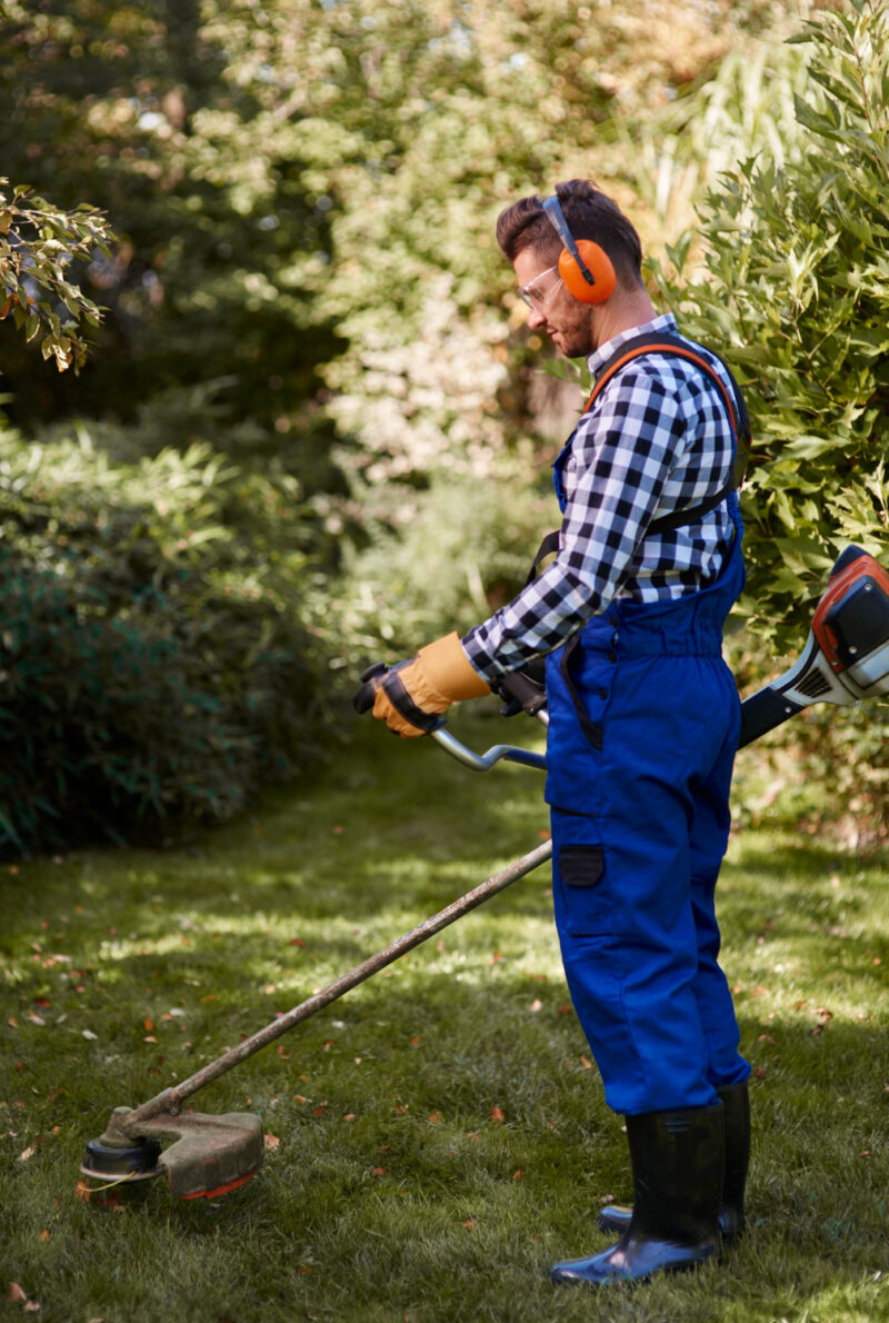 Homme en combinaison bleue utilisant un coupe-bordures dans un jardin, équipé de protège-oreilles et de lunettes de sécurité
