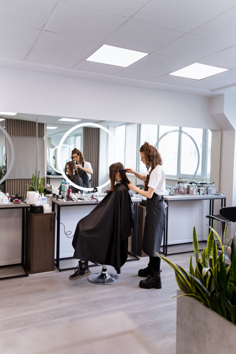 Une coiffeuse coiffe les cheveux d'une cliente assise dans un salon de coiffure moderne avec des miroirs ronds lumineux