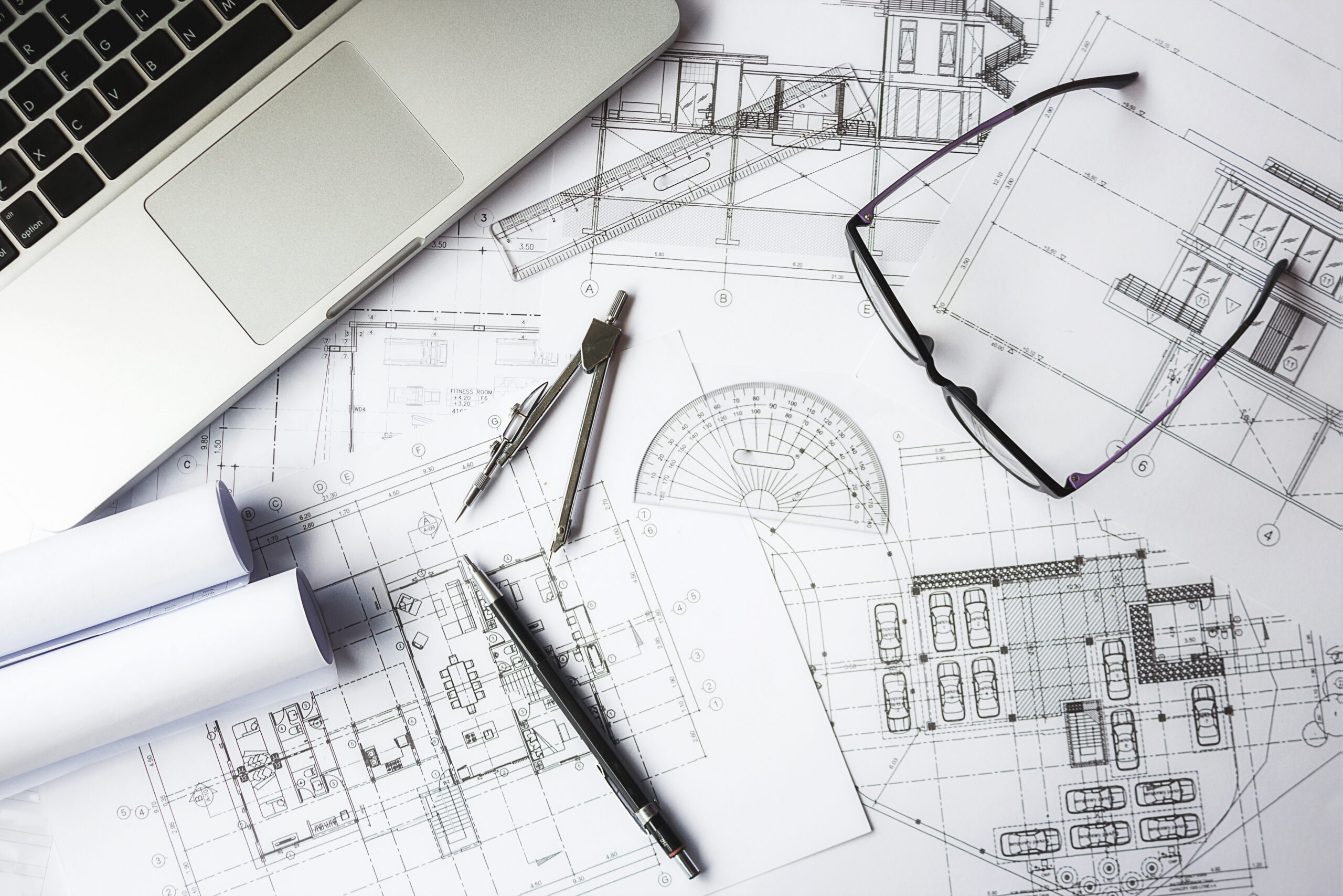 Plans architecturaux étalés sur un bureau avec un compas, un rapporteur, un stylo, des lunettes et un ordinateur portable