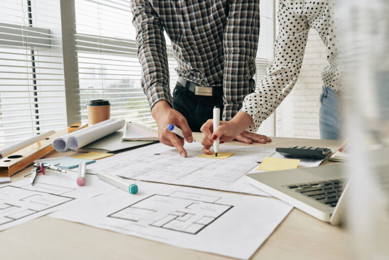 Deux personnes examinant des plans architecturaux étalés sur un bureau avec des outils de dessin et un ordinateur portable