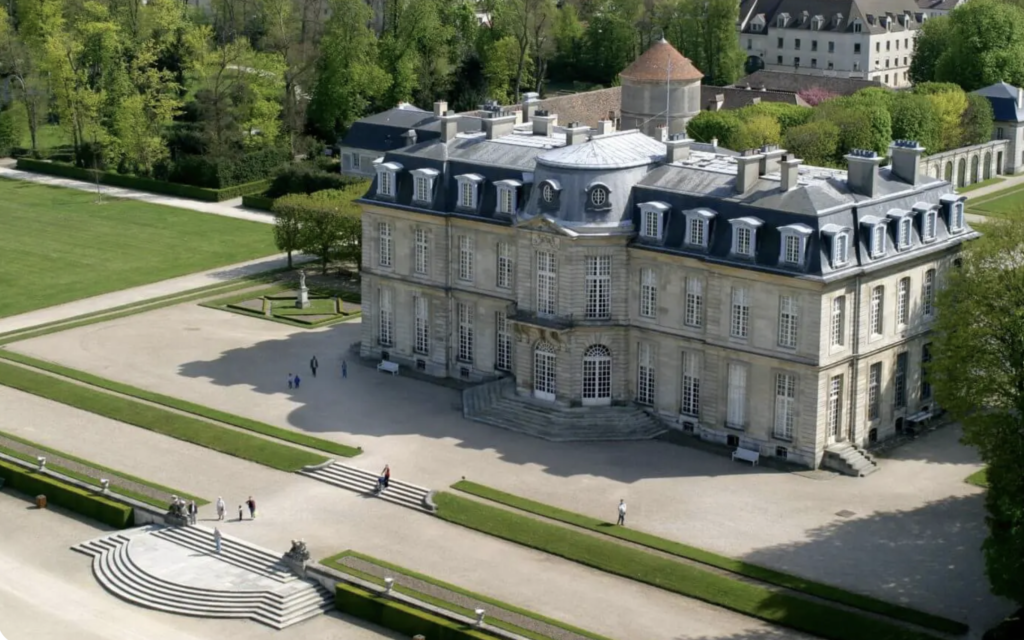 Vue aérienne d'un château classique français entouré de jardins à la française et d'arbres, avec des visiteurs dans la cour