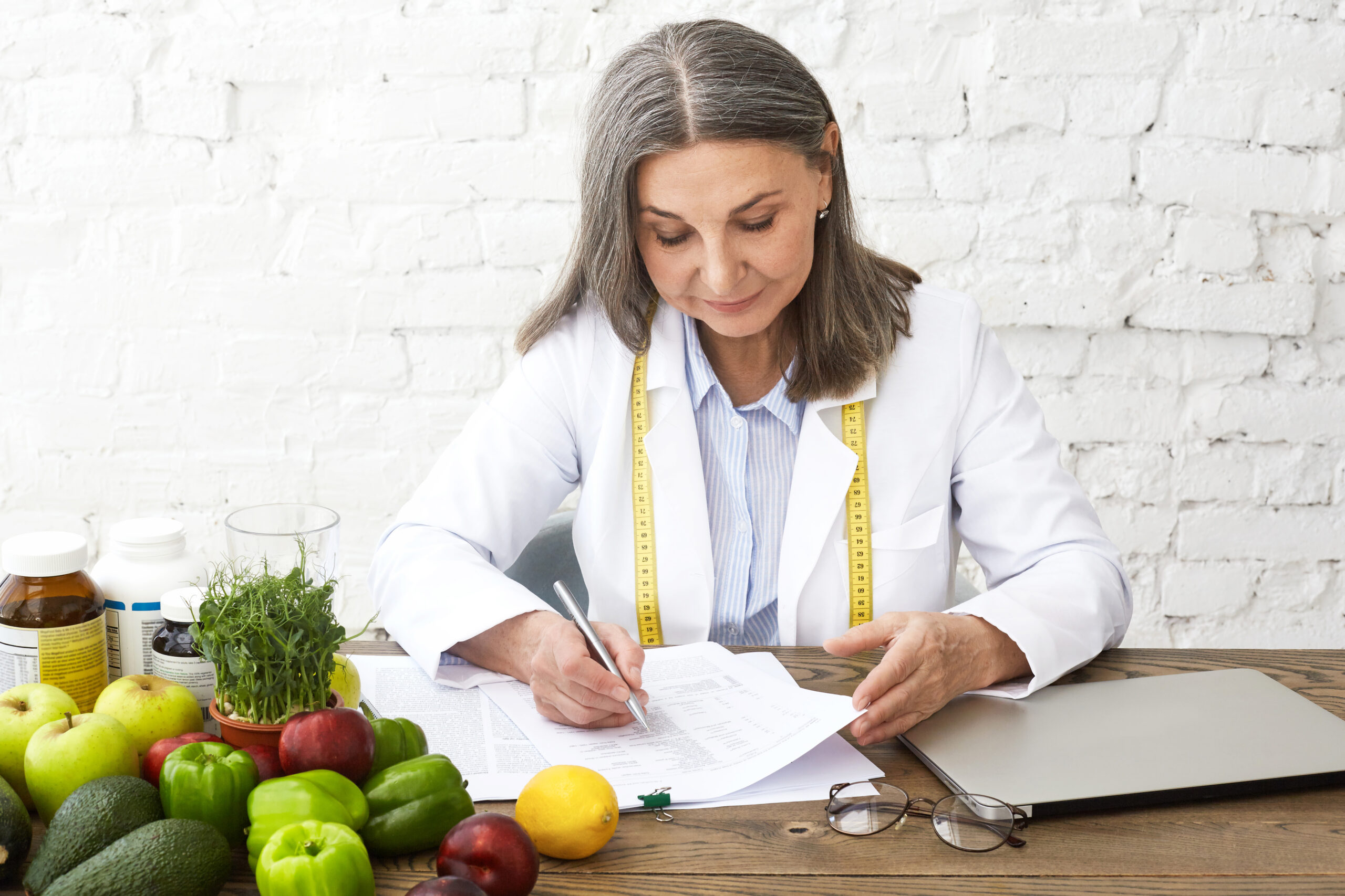 Nutritionniste en blouse blanche avec un mètre ruban, écrivant des notes entourée de fruits, légumes et compléments alimentaires