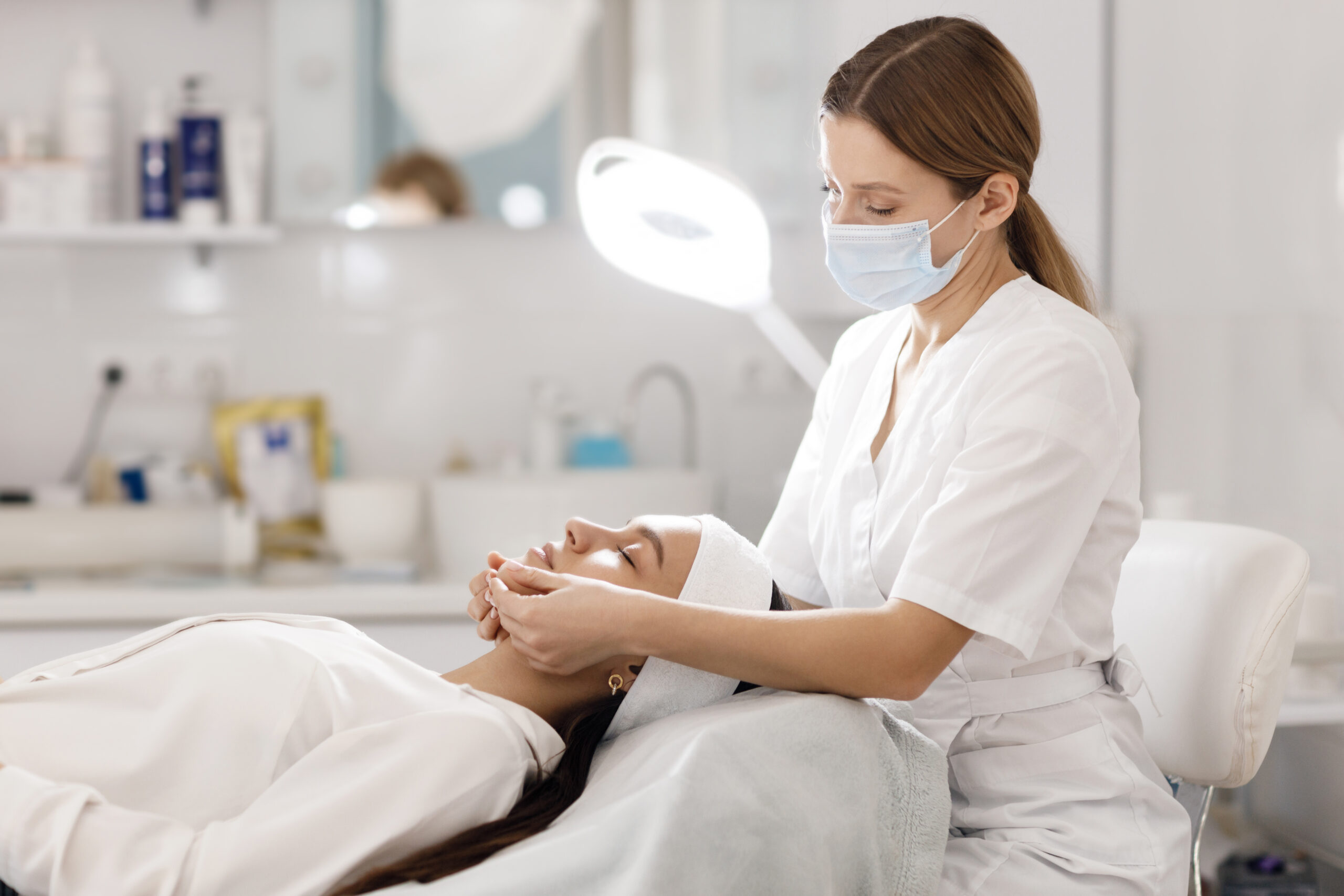 Esthéticienne masquée effectuant un soin du visage sur une cliente allongée dans un institut de beauté