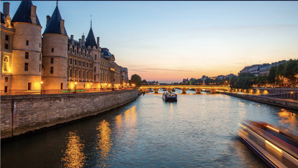 La Conciergerie illuminée au coucher du soleil au bord de la Seine, avec des bateaux naviguant sous un pont parisien