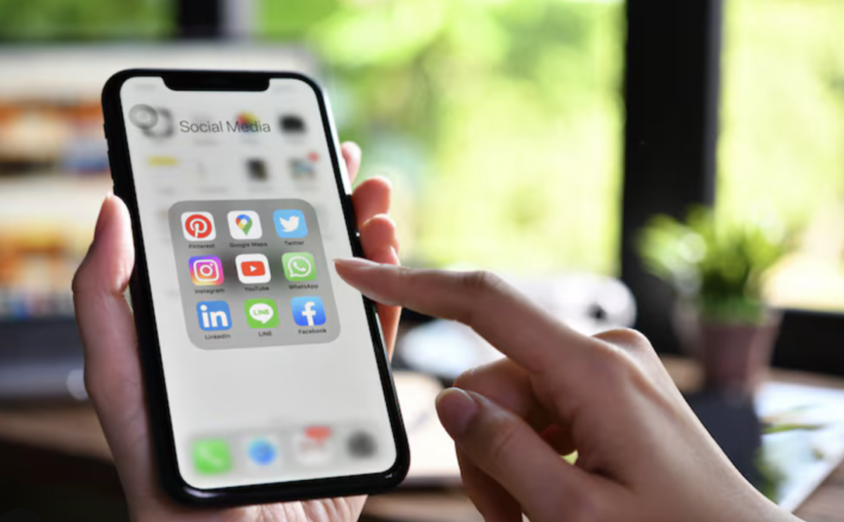 Mains tenant un smartphone affichant un dossier d'applications de réseaux sociaux : Pinterest, Instagram, LinkedIn, YouTube, WhatsApp et Facebook