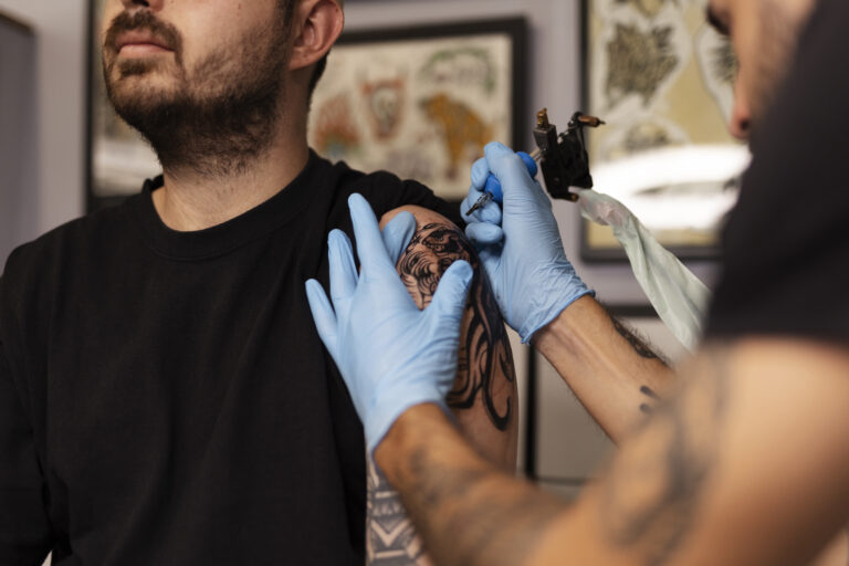 Un tatoueur portant des gants bleus réalise un tatouage sur l'épaule d'un homme dans un salon de tatouage