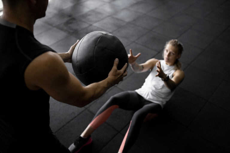 Deux personnes s'entraînent en salle de sport avec un medicine ball, l'une au sol prête à attraper le ballon