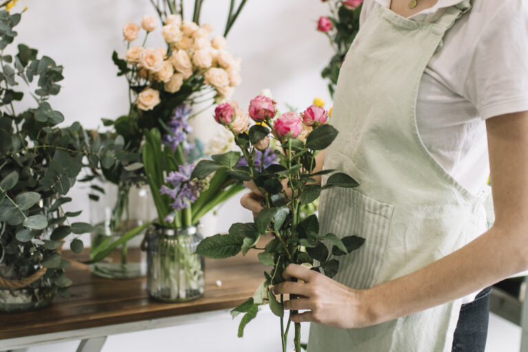 Une fleuriste en tablier vert tient un bouquet de roses roses dans un atelier entouré de fleurs variées