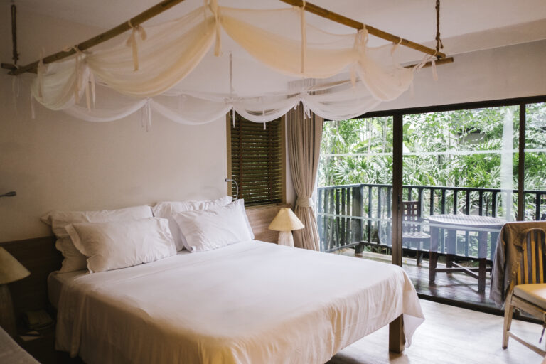 Chambre avec lit baldaquin en bambou, draps blancs et balcon donnant sur une végétation tropicale luxuriante