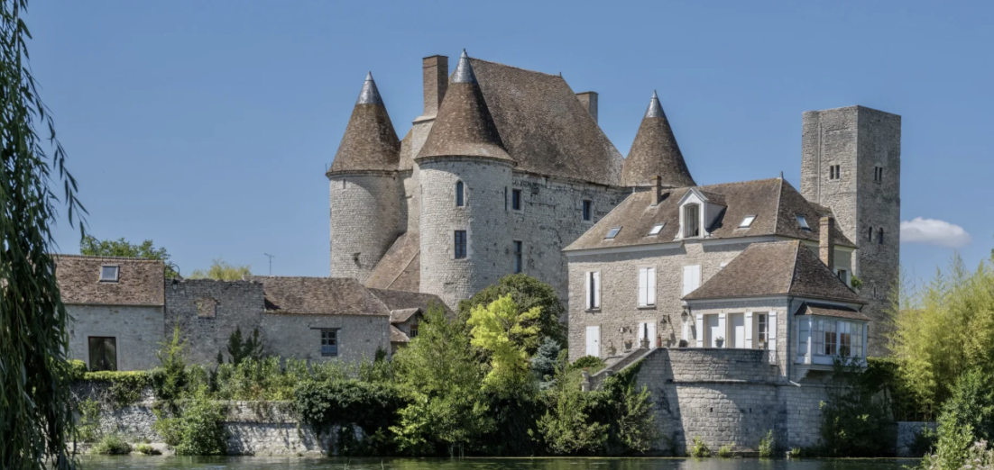 Château médiéval en pierre avec tours rondes et toits en tuiles, entouré de verdure et d'un plan d'eau au premier plan