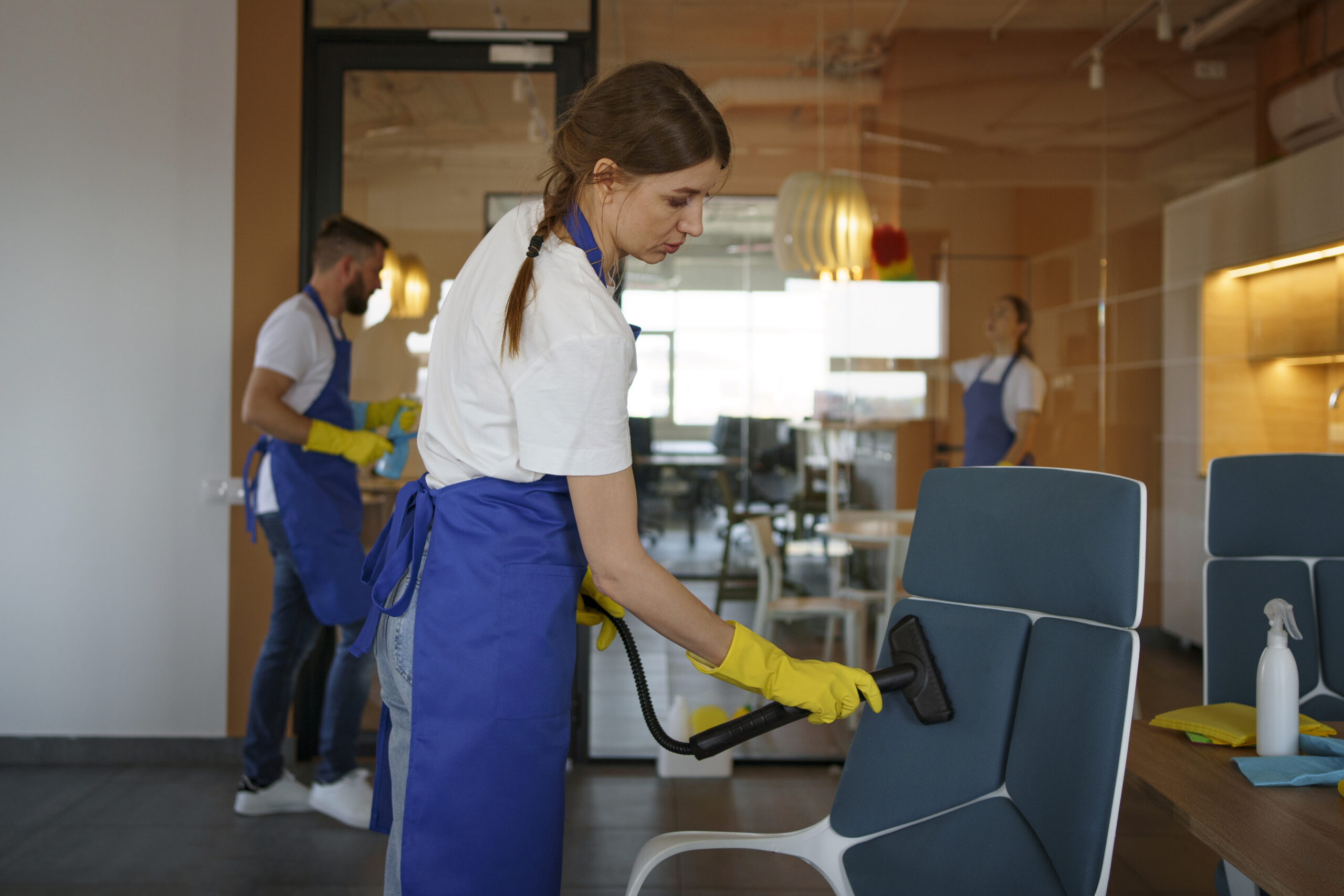 Équipe de nettoyage professionnelle en tabliers bleus nettoyant un bureau, l'une utilisant un nettoyeur vapeur sur une chaise