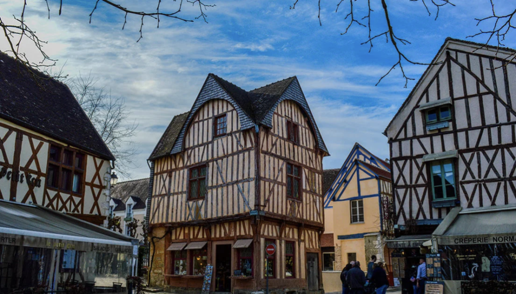 Place médiévale avec maisons à colombages normandes et une crêperie, sous un ciel bleu nuageux