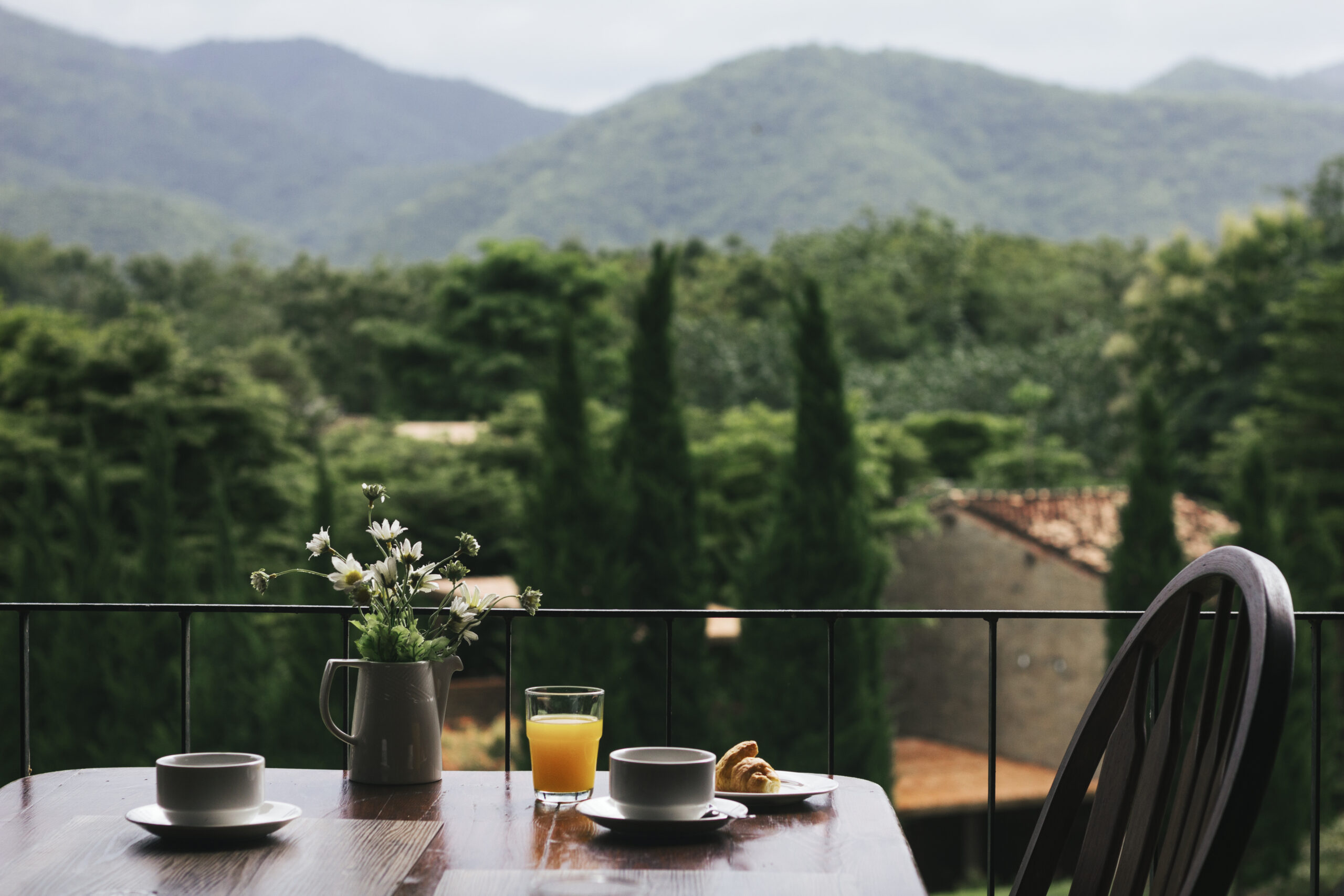 Petit-déjeuner sur une terrasse avec tasses, jus d'orange, croissant et fleurs, vue sur collines verdoyantes et cyprès