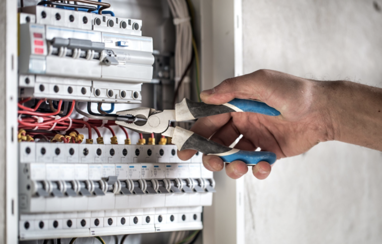 Une main tenant une pince travaillant sur un tableau électrique avec disjoncteurs et câbles multicolores