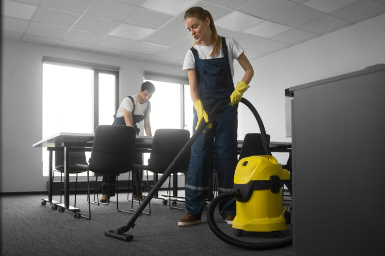 Deux agents d'entretien en salopette bleue nettoient un bureau : une femme passe l'aspirateur, un homme nettoie une table de réunion