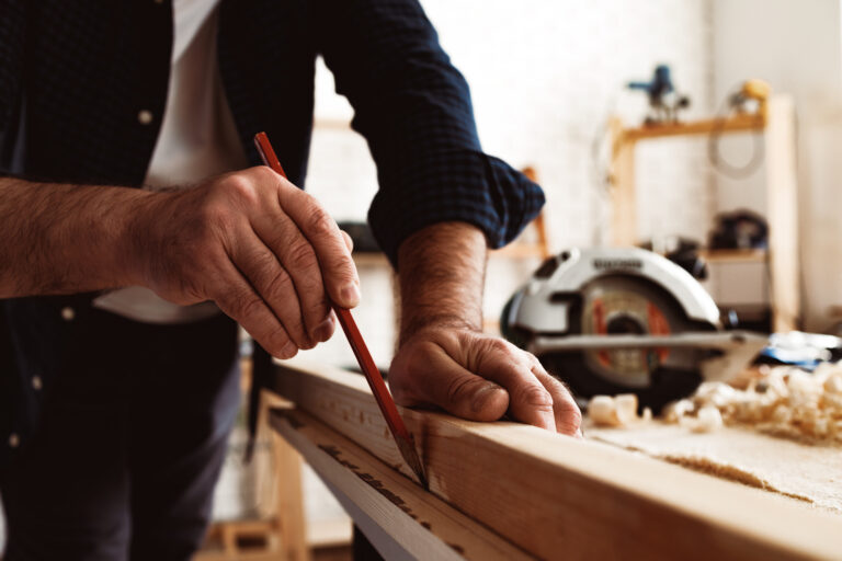 Menuisier traçant une marque au crayon sur une planche en bois dans un atelier, avec une scie circulaire en arrière-plan