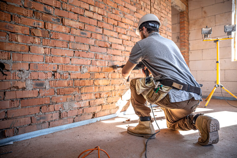 Ouvrier du bâtiment avec casque et ceinture à outils perçant un mur en briques sur un chantier de rénovation